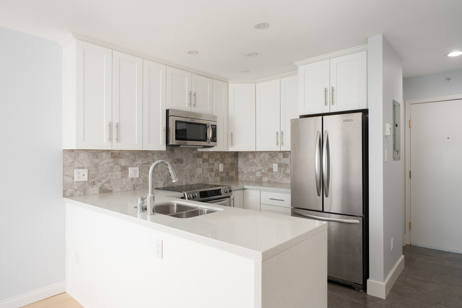 Modern kitchen with white cabinets, stainless steel appliances, a double sink, and a light stone backsplash, viewed from a corner of the countertop.