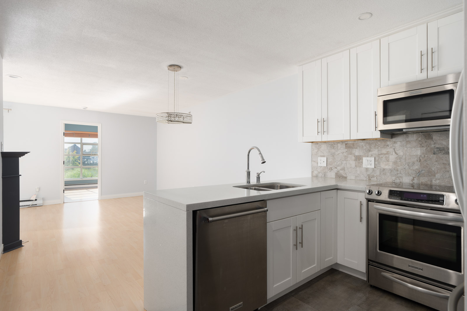 Modern kitchen with white cabinets, stainless steel appliances, and a double sink, opening to a bright, empty living area with light wood flooring and large windows.