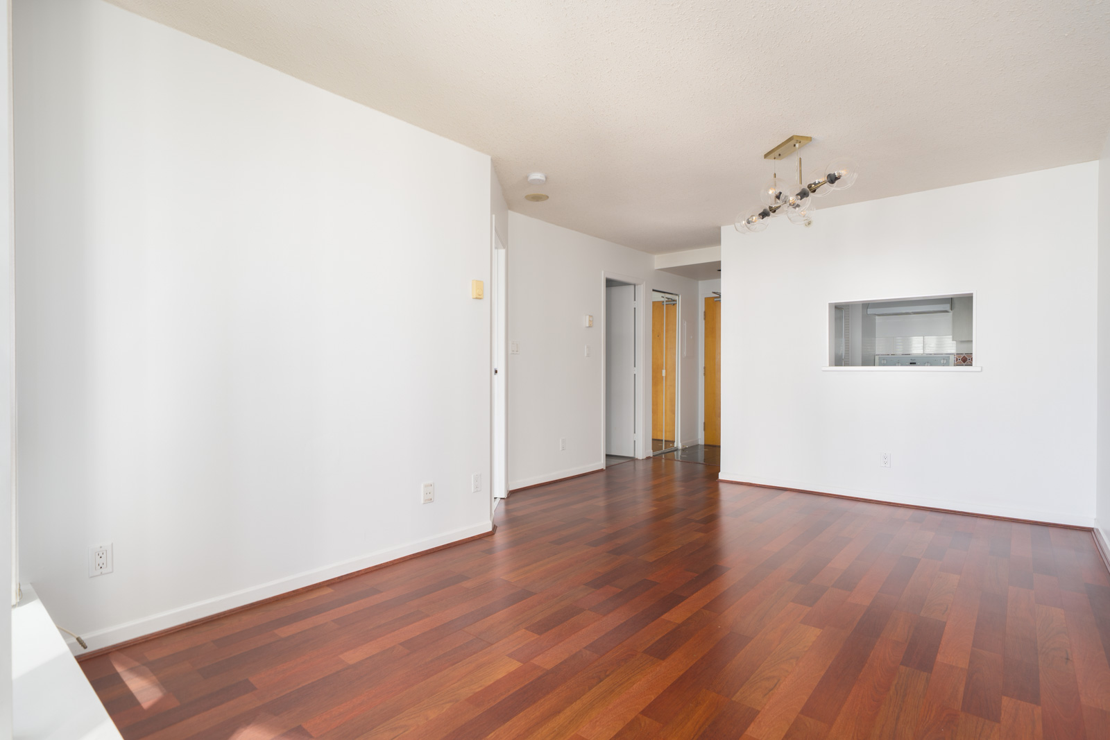 Empty room with white walls, reddish-brown hardwood floors, a modern ceiling light fixture, and a pass-through window to the kitchen.