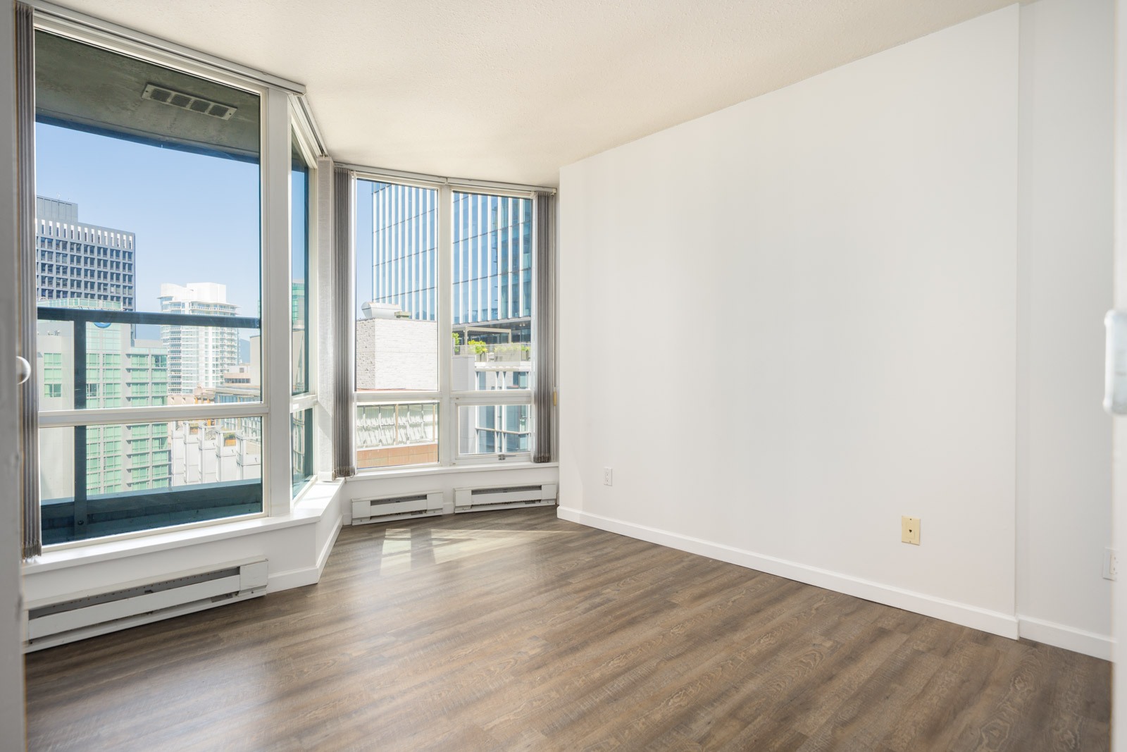 Unfurnished corner apartment room with large windows, white walls, and wood laminate flooring, offering a view of city buildings outside.