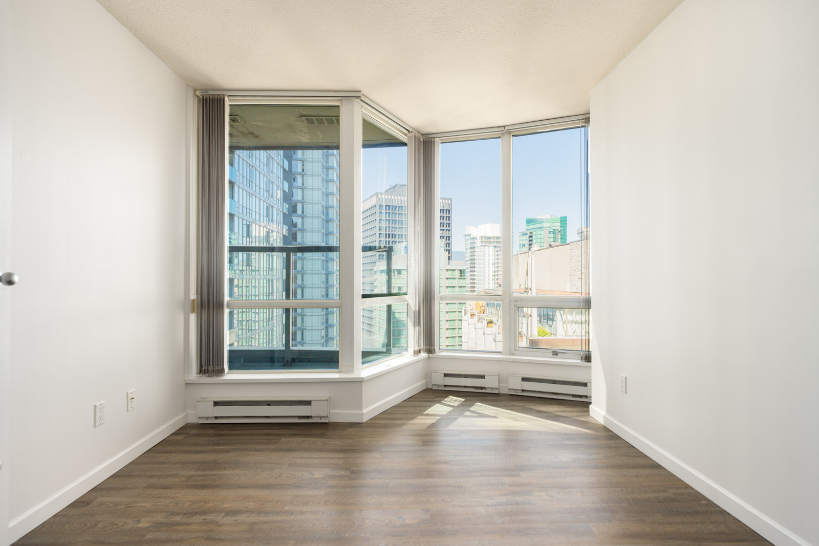 Empty corner room with large floor-to-ceiling windows, white walls, and wood flooring, offering a view of city buildings and a balcony outside.