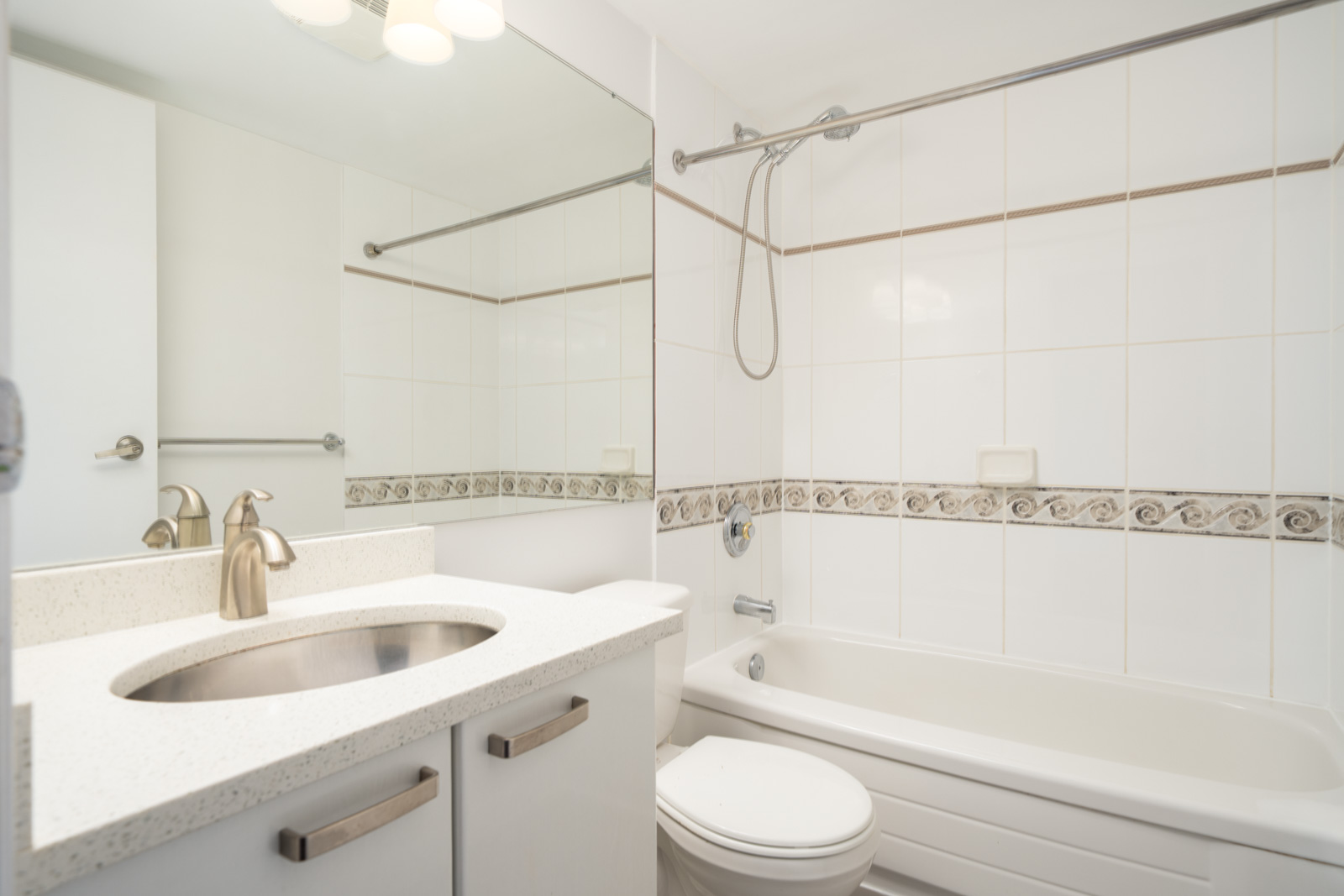 Modern bathroom with a white vanity, sink, mirror, toilet, and bathtub with a showerhead. White tile walls feature a decorative border. Bright, clean, and minimal design.