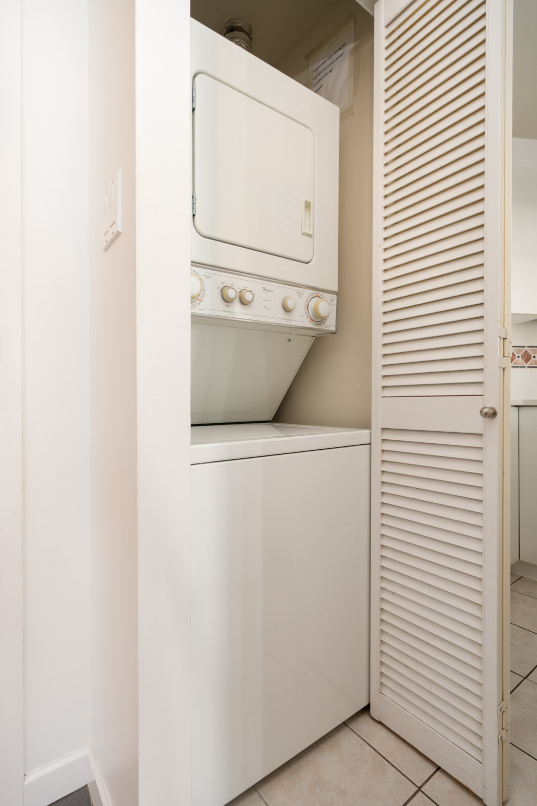 A stacked washer and dryer unit is installed in a small laundry closet with a partially open louvered door in a tiled room.