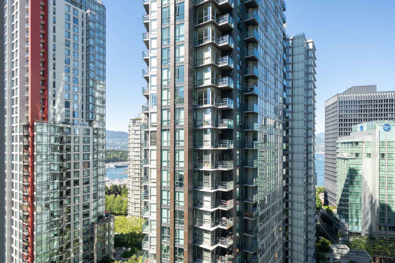 High-rise residential and office buildings with glass facades stand close together in an urban downtown area, with water and mountains visible in the background.