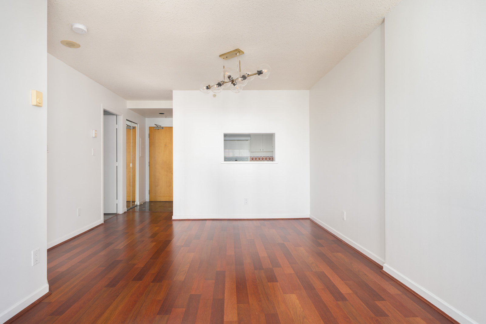 Empty room with white walls, wooden flooring, modern ceiling light fixture, and a small interior window opening to another room.
