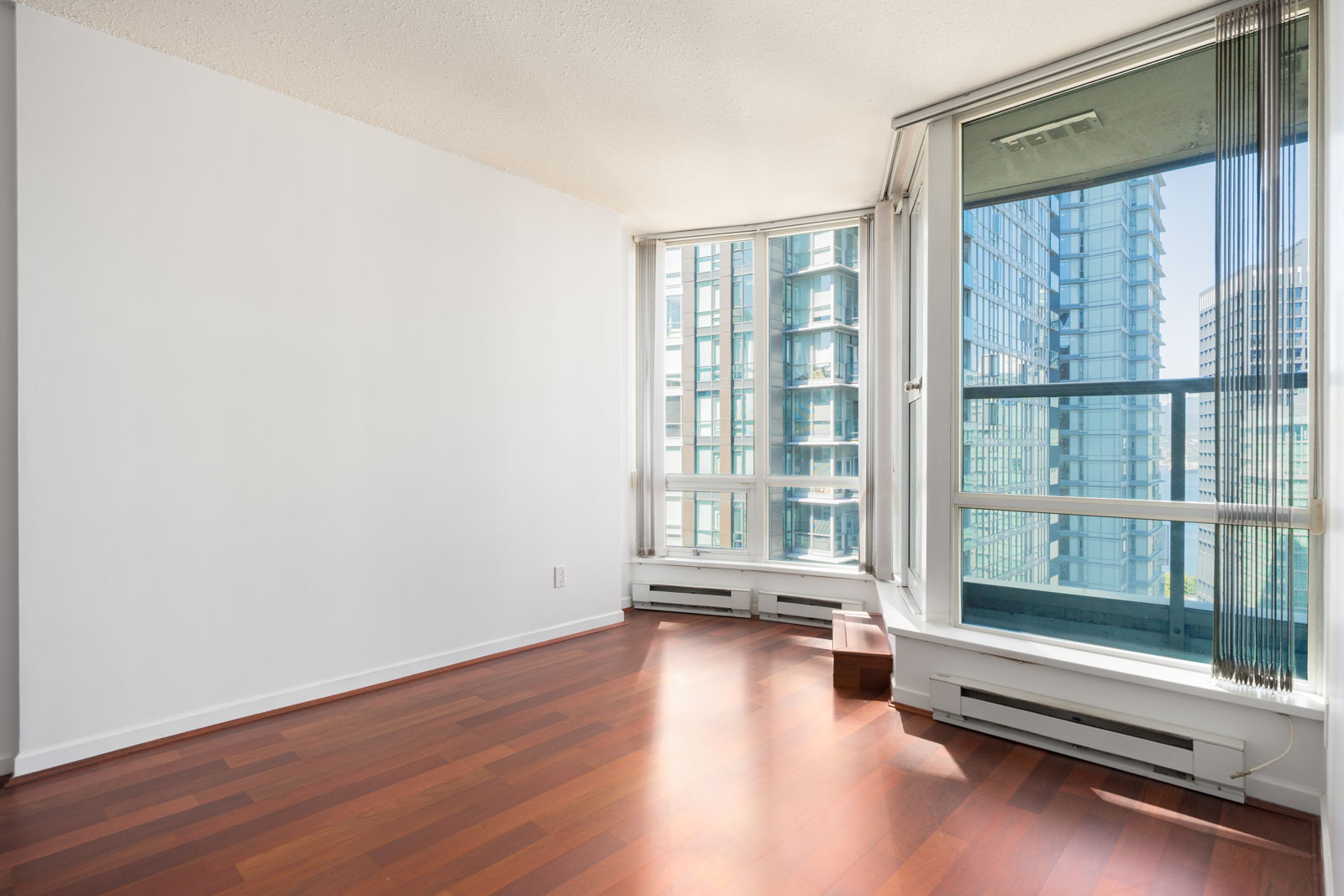 Bright, empty room with large windows, white walls, and polished wood floors overlooking modern high-rise buildings.