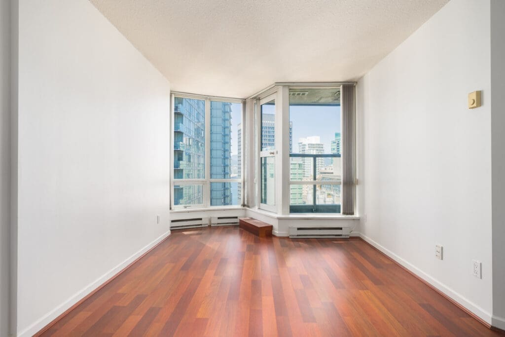 Unfurnished room with hardwood floors, white walls, large corner windows, and a glass door overlooking modern city buildings.
