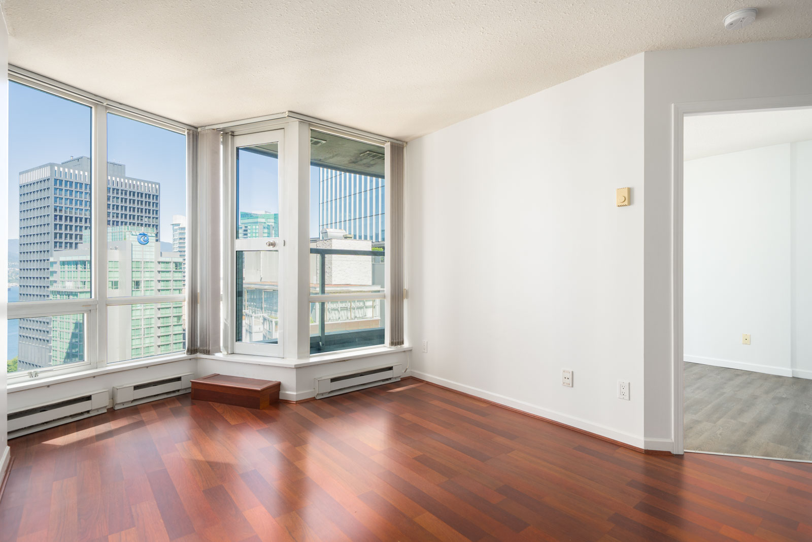 Bright corner room with large windows, hardwood floors, white walls, and a partial city view; an open doorway leads to an adjacent room.