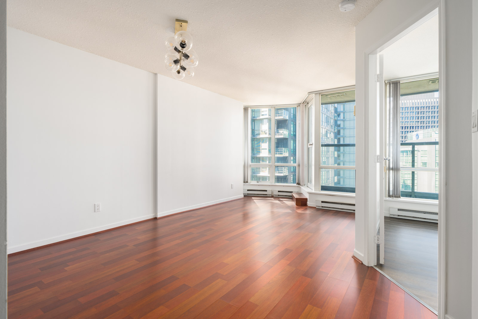 Empty modern apartment room with reddish wood flooring, white walls, large windows, a ceiling light fixture, and an open door leading to another area.