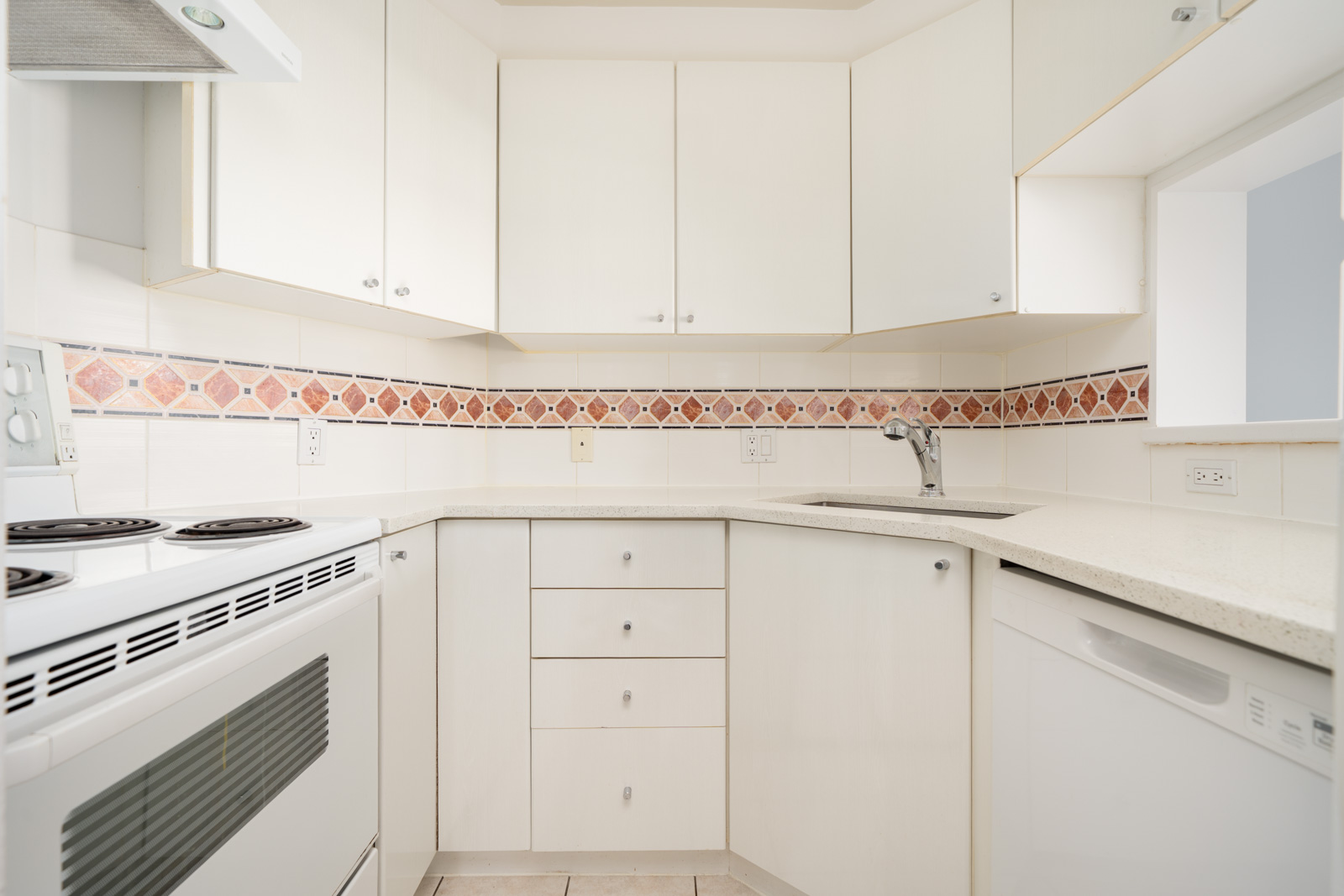 Small, white kitchen with electric stove, dishwasher, sink, and upper and lower cabinets. Light-colored countertops and decorative tile backsplash featuring a red and beige geometric pattern.