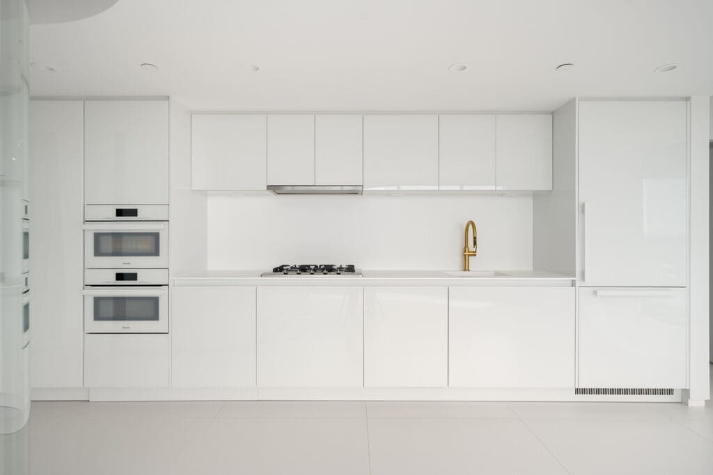 Minimalist modern white kitchen with built-in oven, stove, gold faucet, and sleek cabinets in a clean, bright setting.