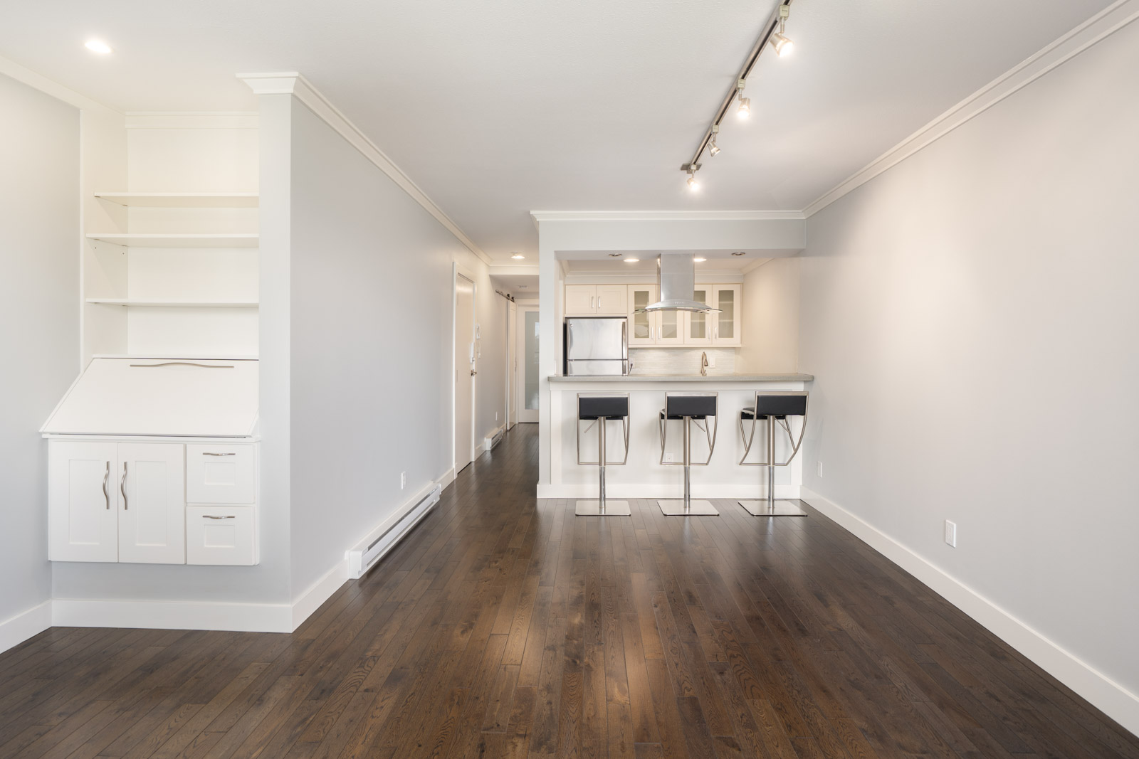 Modern, unfurnished open-concept kitchen and living area with dark wood flooring, white cabinets, and three bar stools at a breakfast bar.