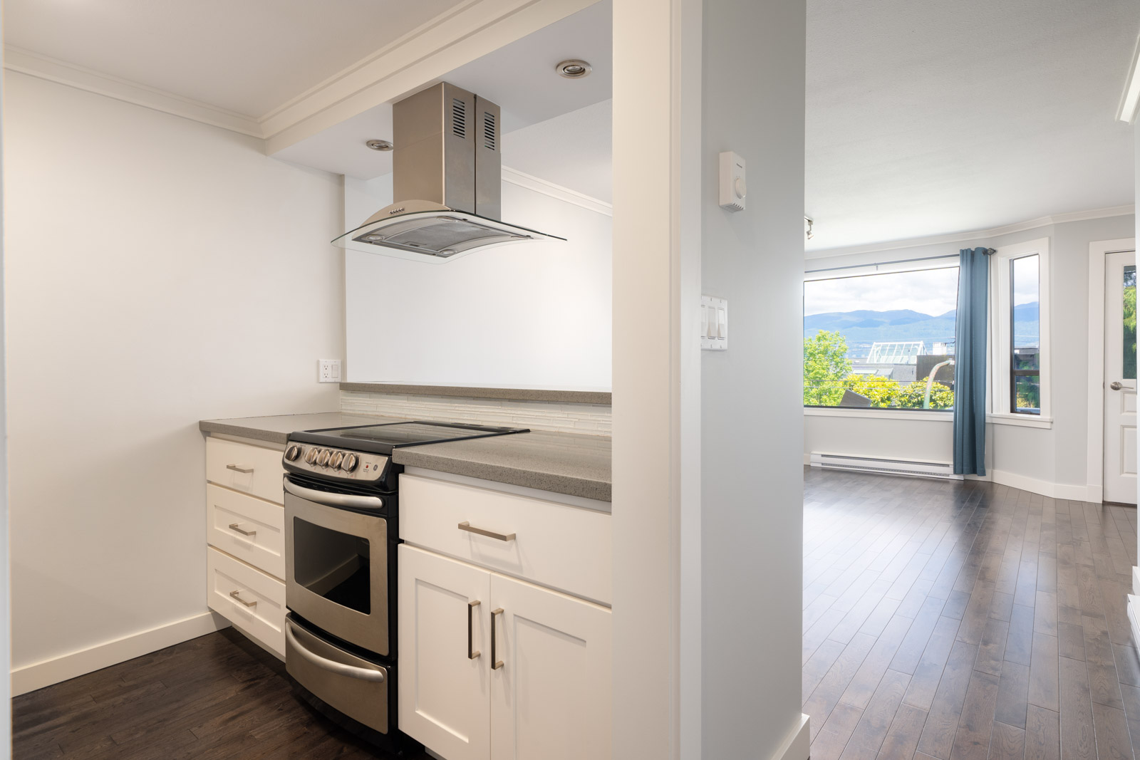 Modern kitchen with white cabinets and stainless steel stove adjacent to a bright living area with large windows and dark wood flooring.