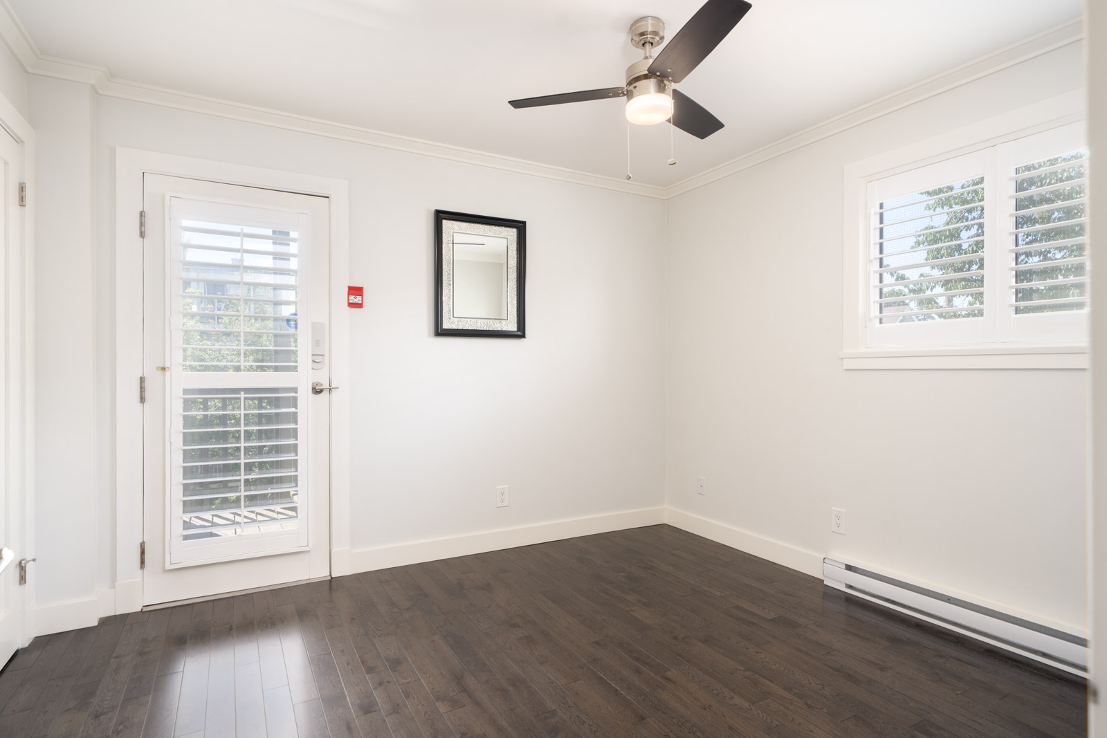 Empty room with dark wood floor, white walls, ceiling fan, window with shutters, wall mirror, and a door with glass panels and shutters leading outside.