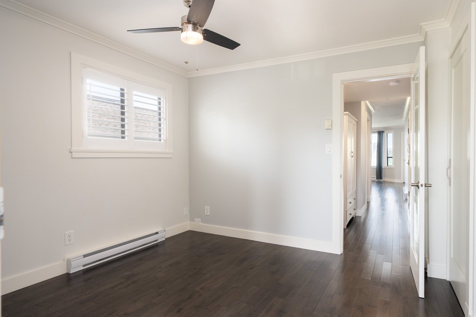 Empty, unfurnished room with light gray walls, dark wood floor, ceiling fan, window with white shutters, and an open door leading to a hallway.