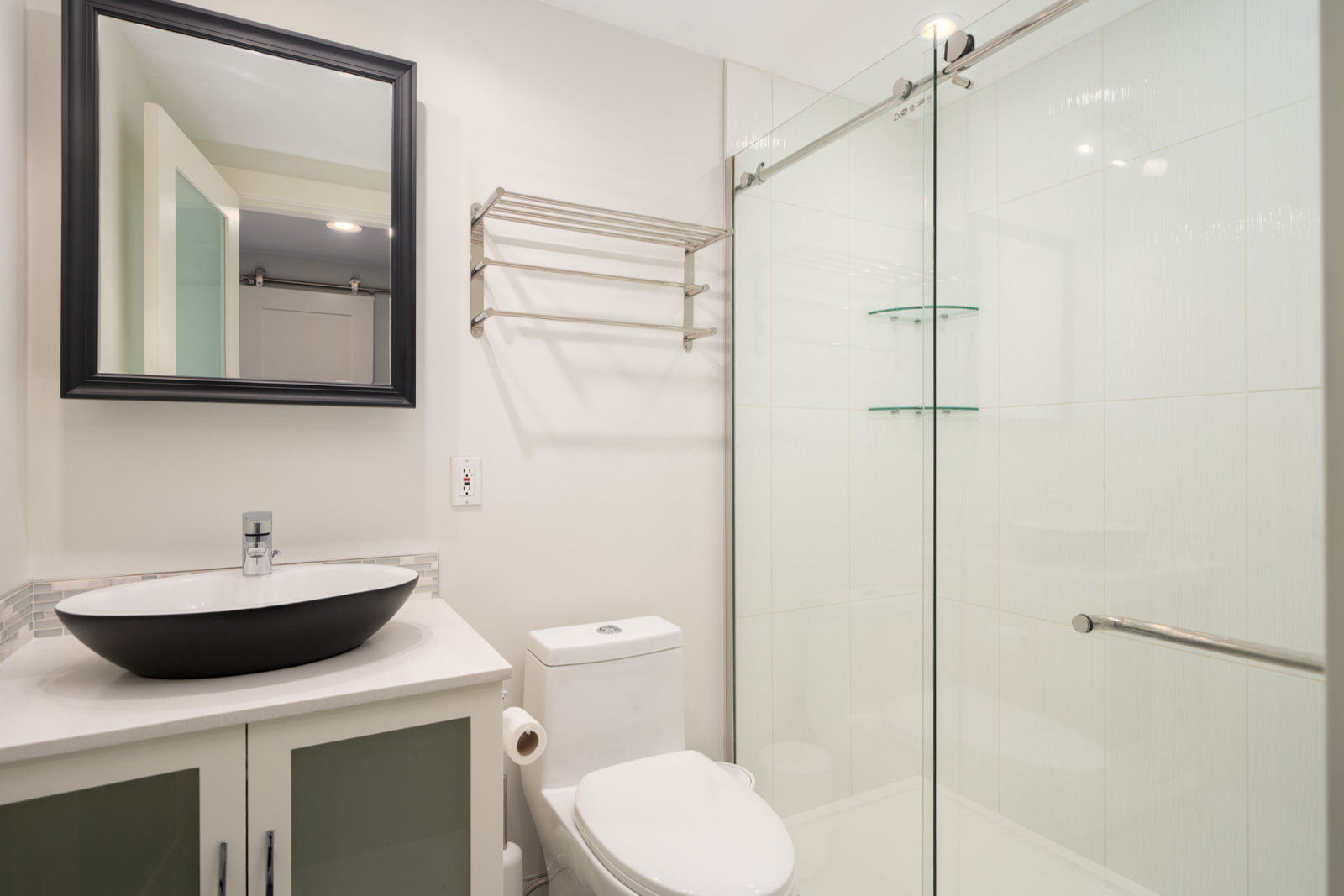 Modern bathroom with a vessel sink, wall-mounted mirror, glass shower enclosure, towel rack, white toilet, and light-colored walls.