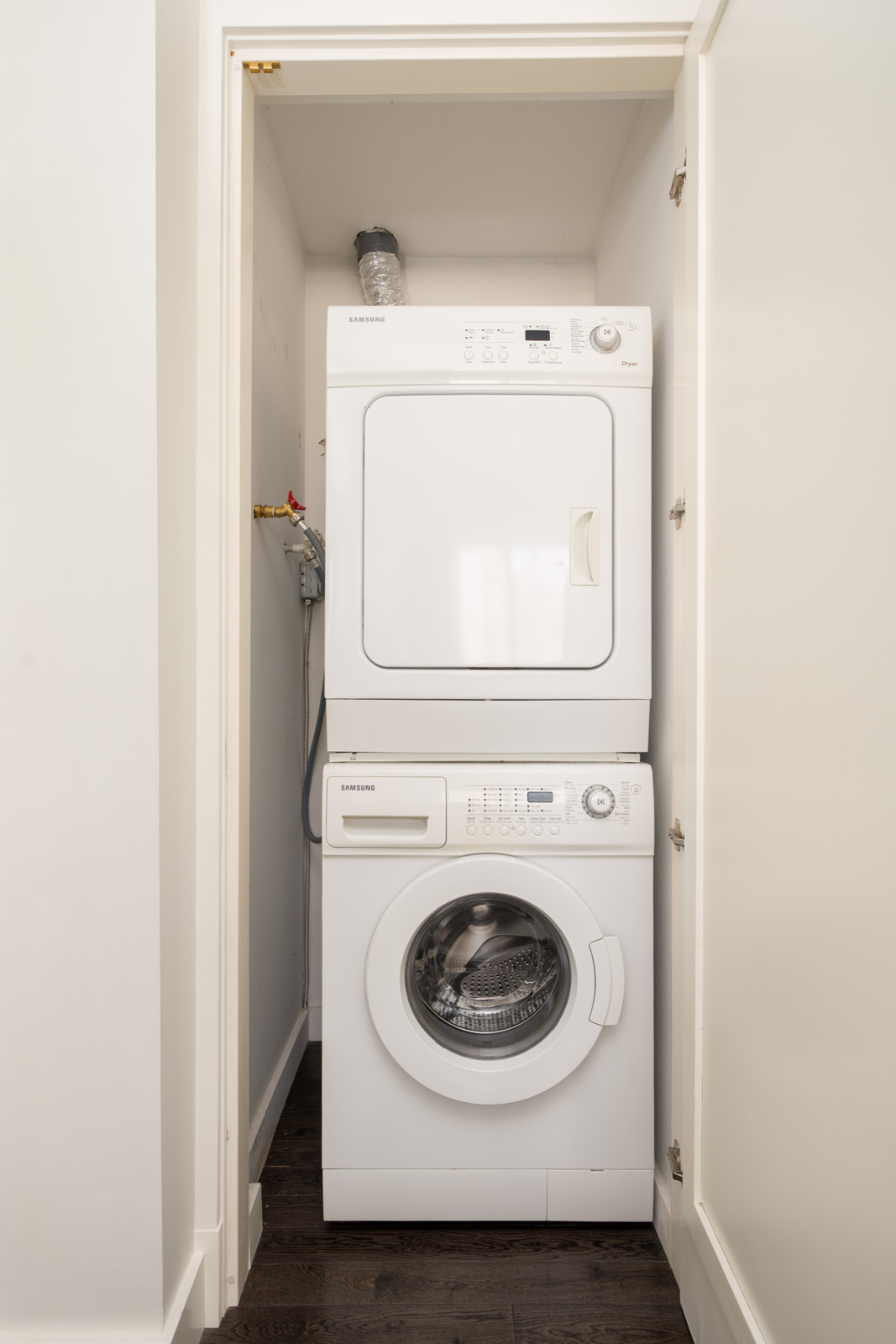 Stacked white washer and dryer units in a small laundry closet with white walls and a dark wood floor.