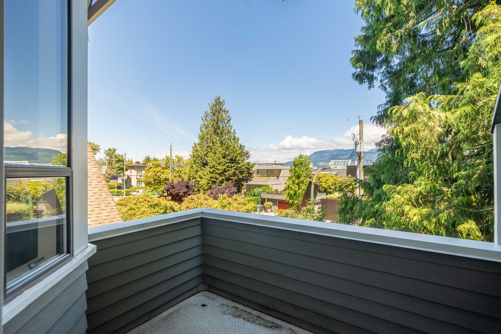 A small balcony with gray siding overlooks a residential neighborhood with trees and houses under a clear blue sky.