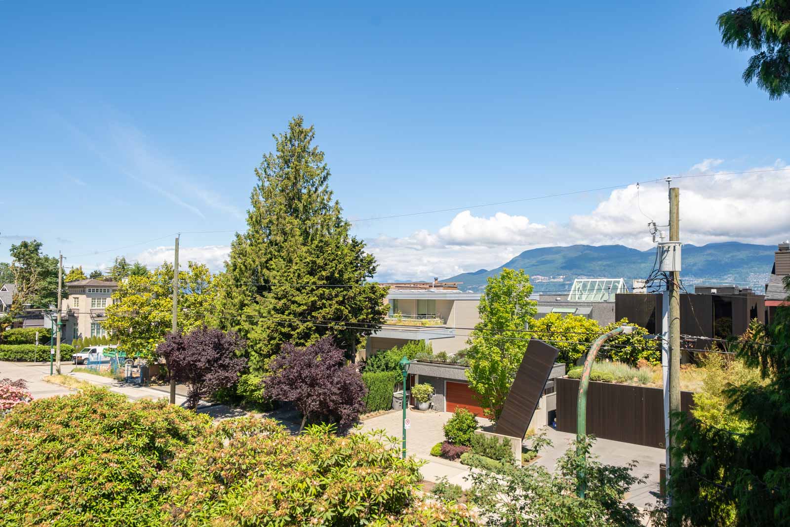 Residential neighborhood with trees, modern houses, utility poles, and distant mountains under a clear blue sky.