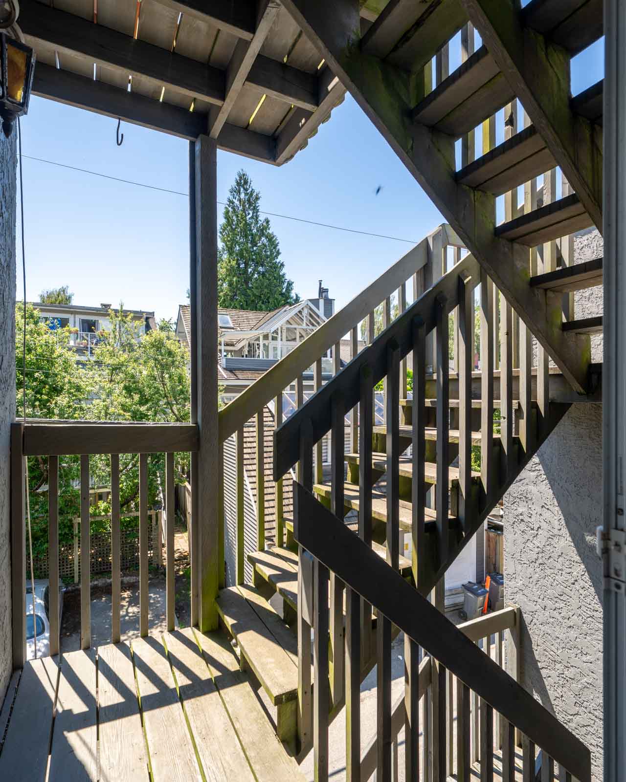 Wooden outdoor staircase with railings and landings, viewed from a shaded area. Sunlight illuminates parts of the stairs, with trees and buildings visible in the background.