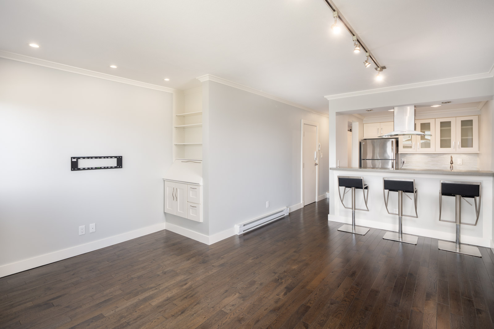 Bright, empty modern living space with dark hardwood floors, white walls, a built-in desk, and a kitchen with a breakfast bar and three stools.