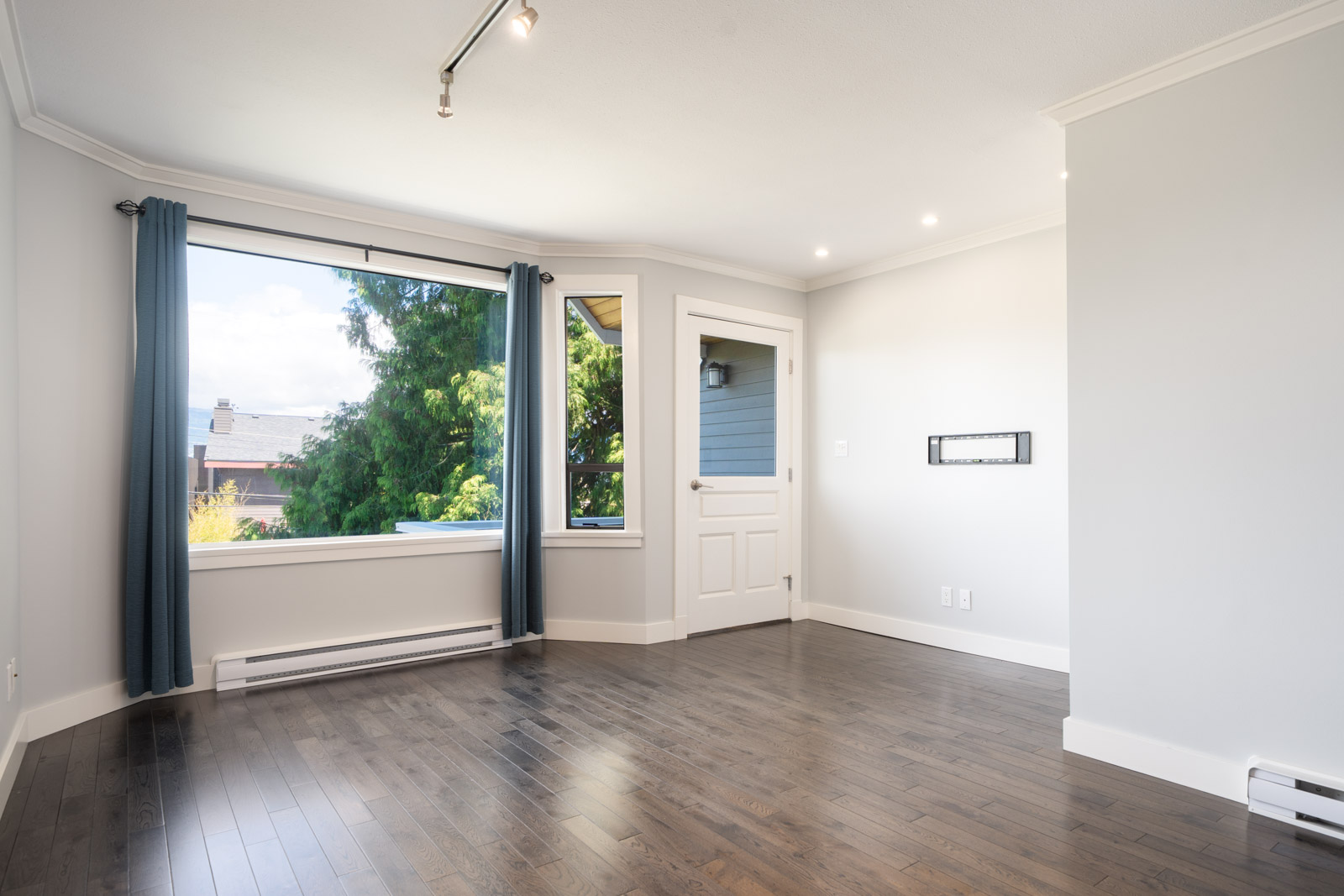 Empty room with light gray walls, dark wood flooring, a large window with blue curtains, a white door, and ceiling track lighting.