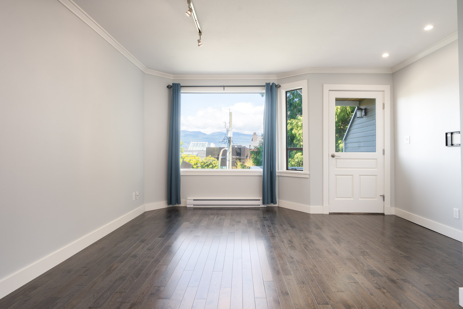Empty room with dark wood floors, white walls, large window with blue curtains, and a white door leading outside; view of mountains and greenery visible through the window.