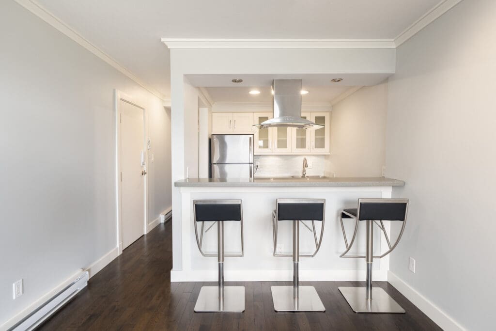 Modern kitchen with stainless steel appliances, white cabinets, and a breakfast bar with three black barstools on dark wood flooring.