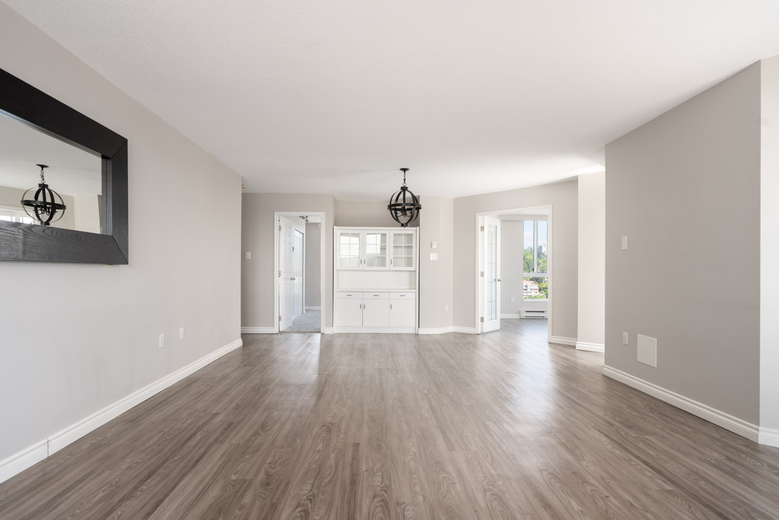 Empty living room with light gray walls, wood flooring, a large wall mirror, ceiling light fixture, and built-in white cabinetry near a doorway leading to a bright adjacent room.