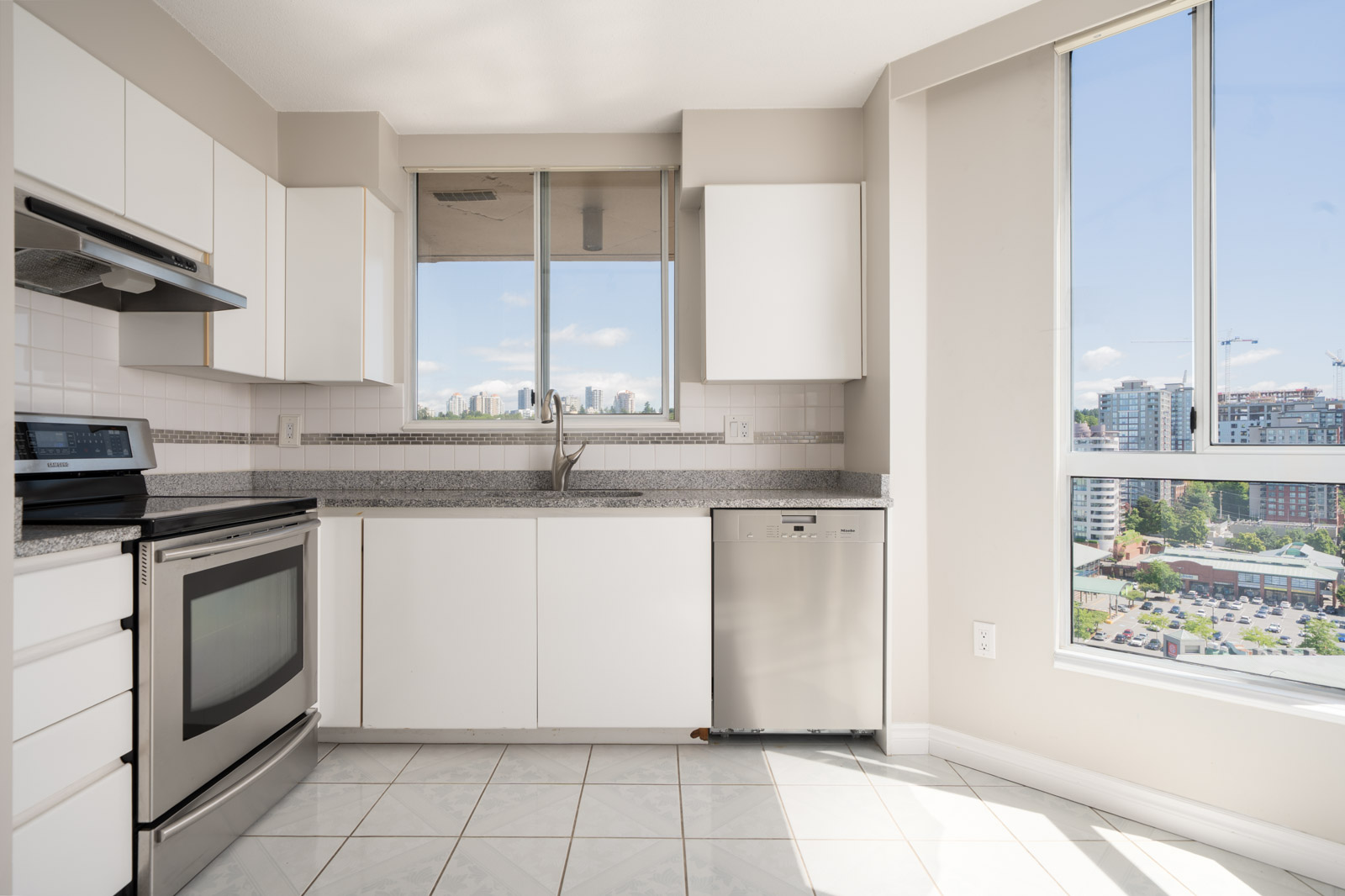 Modern kitchen with white cabinets, stainless steel appliances, tiled floor, and large windows offering a city view.