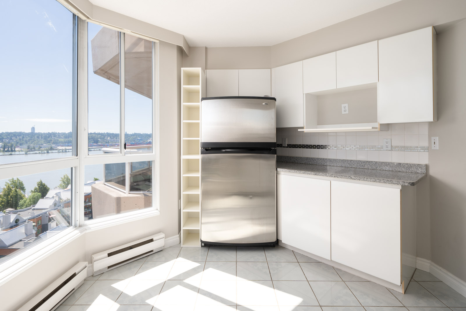 A modern kitchen with white cabinets, a stainless steel refrigerator, granite countertops, tiled floor, and large window overlooking a river and cityscape.
