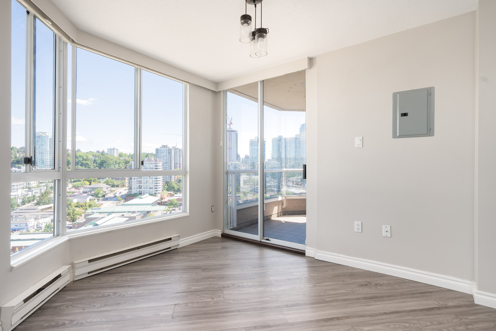 Unfurnished corner apartment room with large windows, light wood flooring, and balcony access overlooking a cityscape on a sunny day.