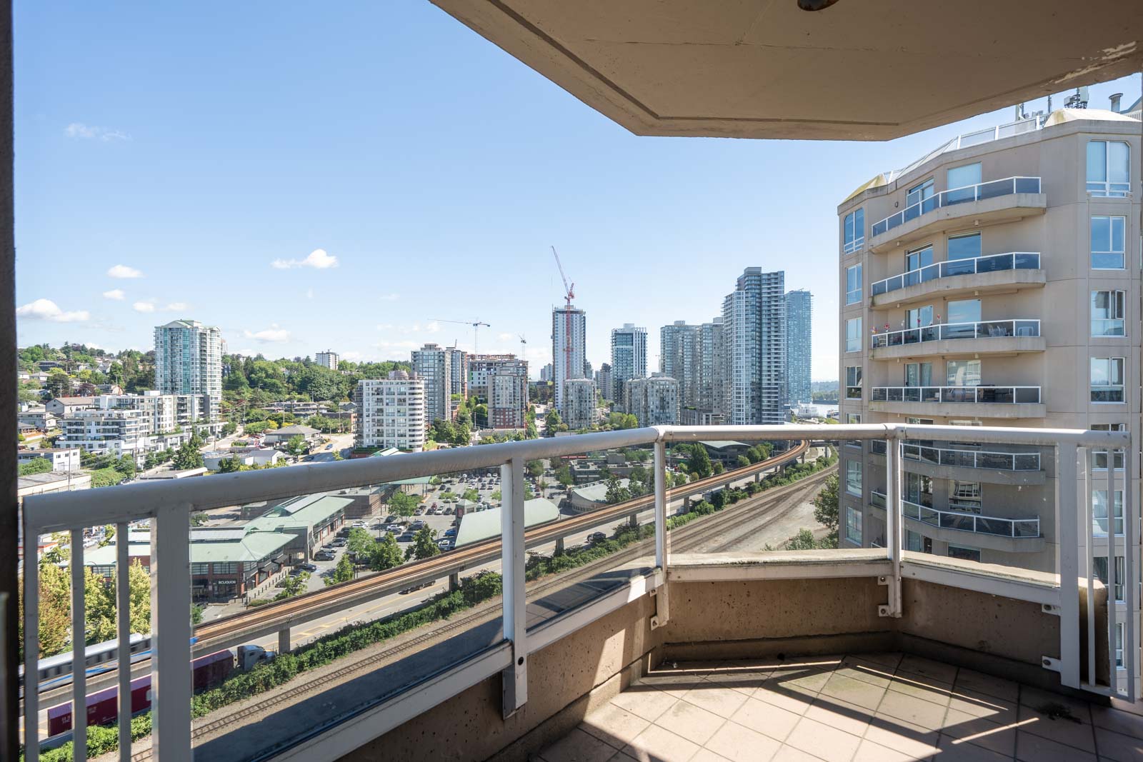 View from a balcony showing nearby rail tracks, mid-rise and high-rise buildings, and a clear sky over an urban cityscape.