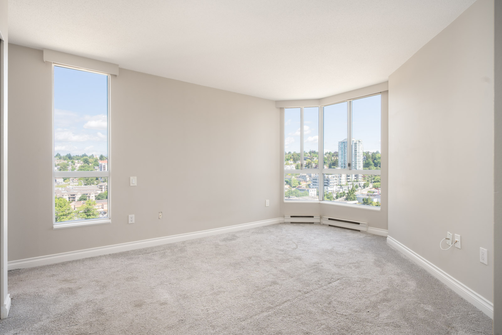 Empty, carpeted room with beige walls, two tall windows, and a corner of floor-to-ceiling windows showing a cityscape and greenery outside.