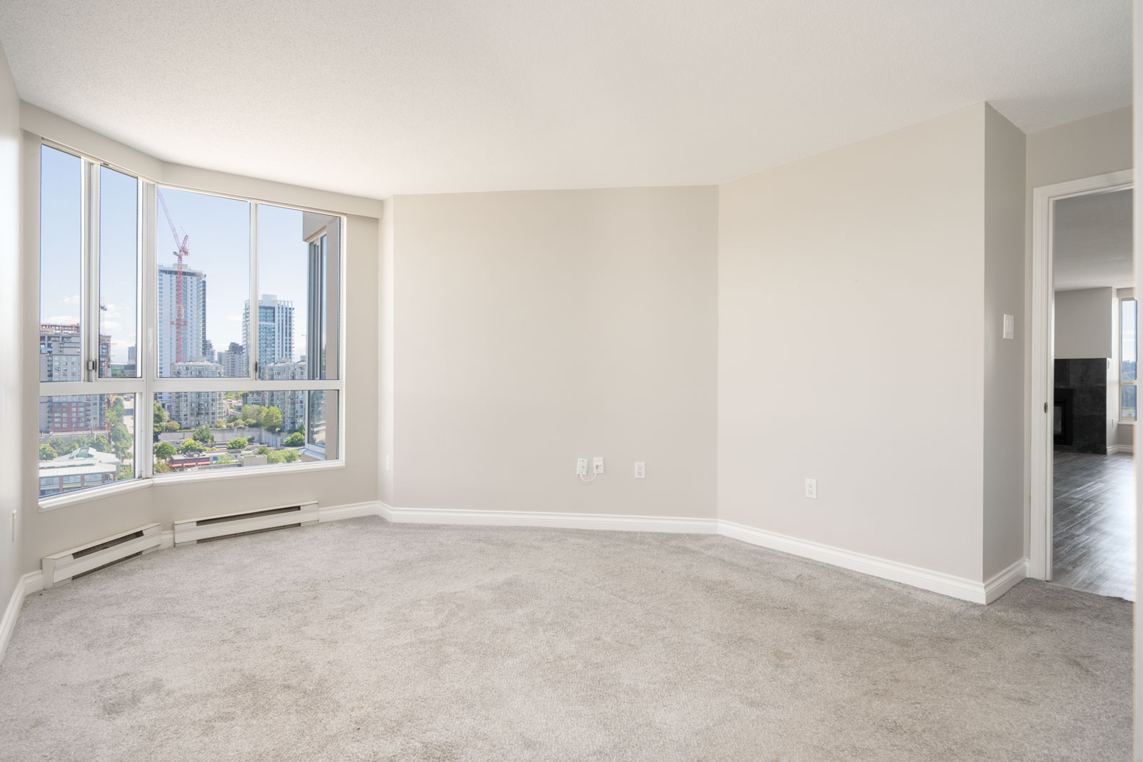 Empty room with light-colored walls and carpet, large bay window showing a city view, and an open doorway leading to another room.
