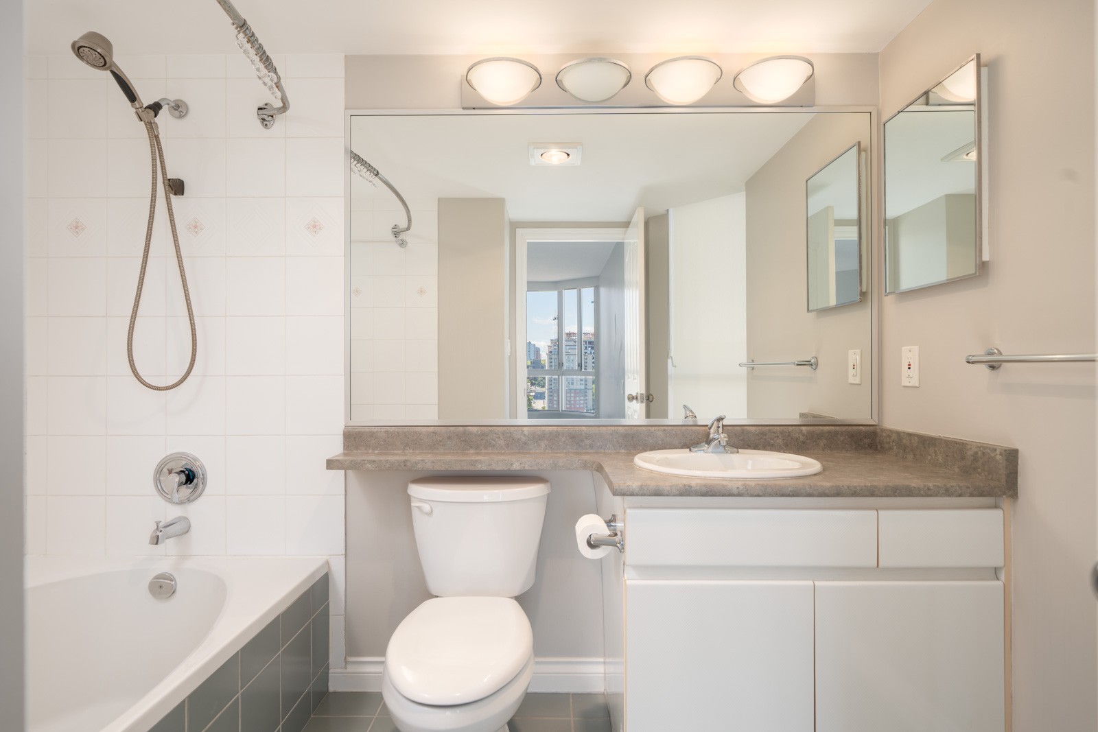 Modern bathroom with a bathtub, showerhead, toilet, large mirror, sink, and white cabinets; cityscape visible through an open doorway.