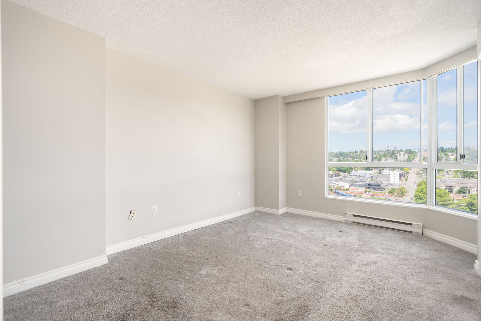 Empty room with light gray carpet, beige walls, baseboard heater, and large windows showing a cityscape and blue sky outside.