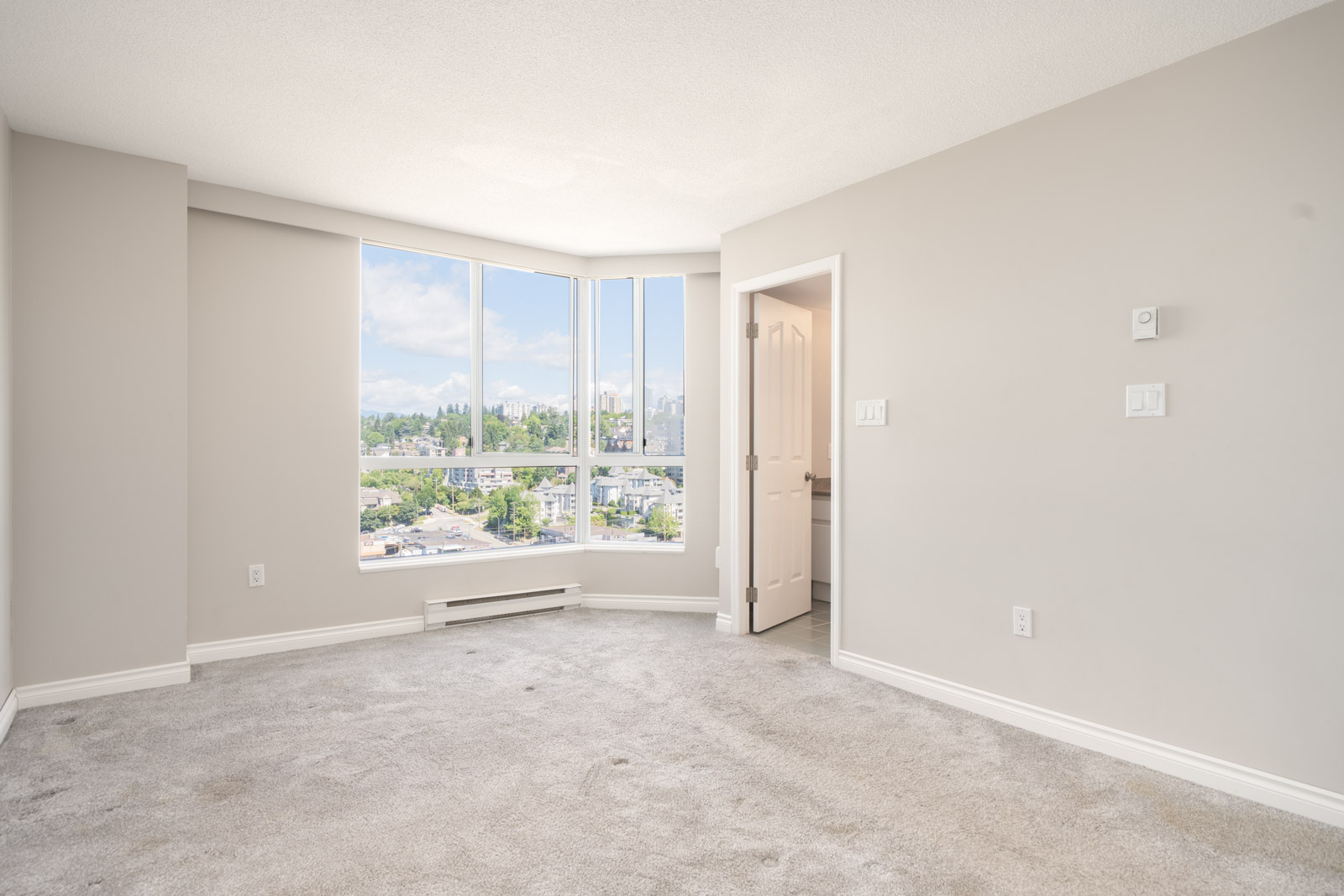 Unfurnished room with light grey carpet, beige walls, large window showing a cityscape, and a door leading to an adjacent room.