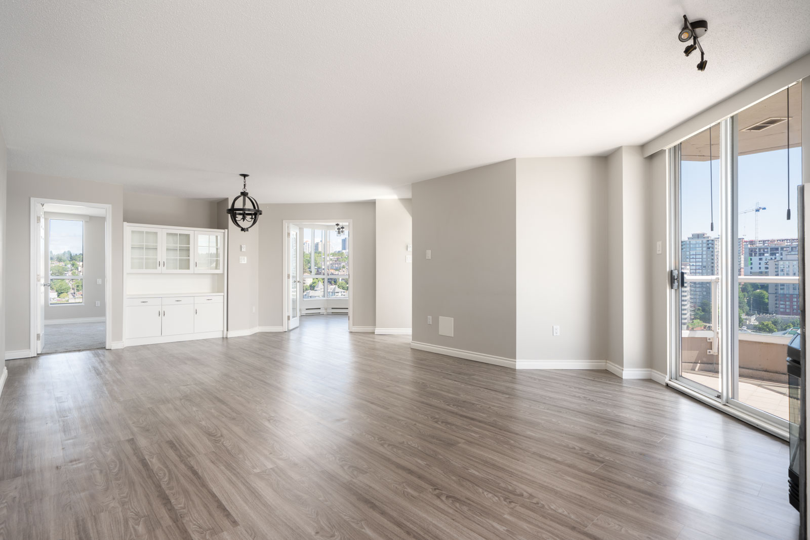 Spacious empty living room with light wood flooring, large windows, sliding glass doors to a balcony, and built-in white cabinetry.