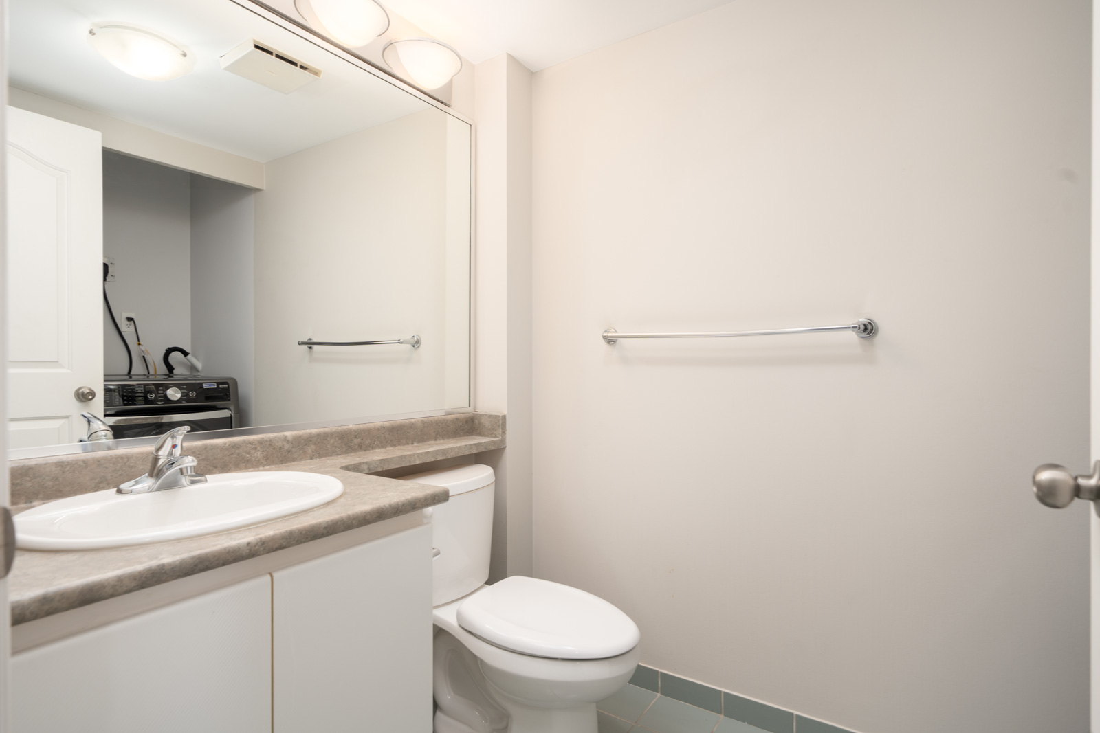 A clean, modern bathroom with a sink, toilet, large mirror, towel bar, and a glimpse of a washer and dryer in an adjacent room.