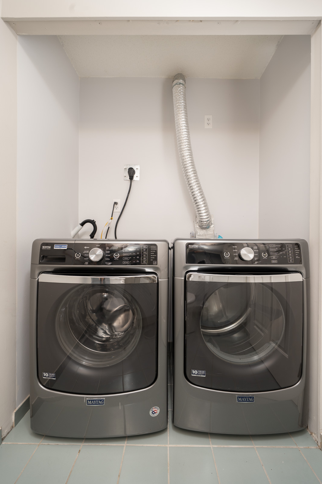 A front-loading washer and dryer set, both silver Maytag models, installed side by side in a laundry alcove with visible vent hose and power outlets.