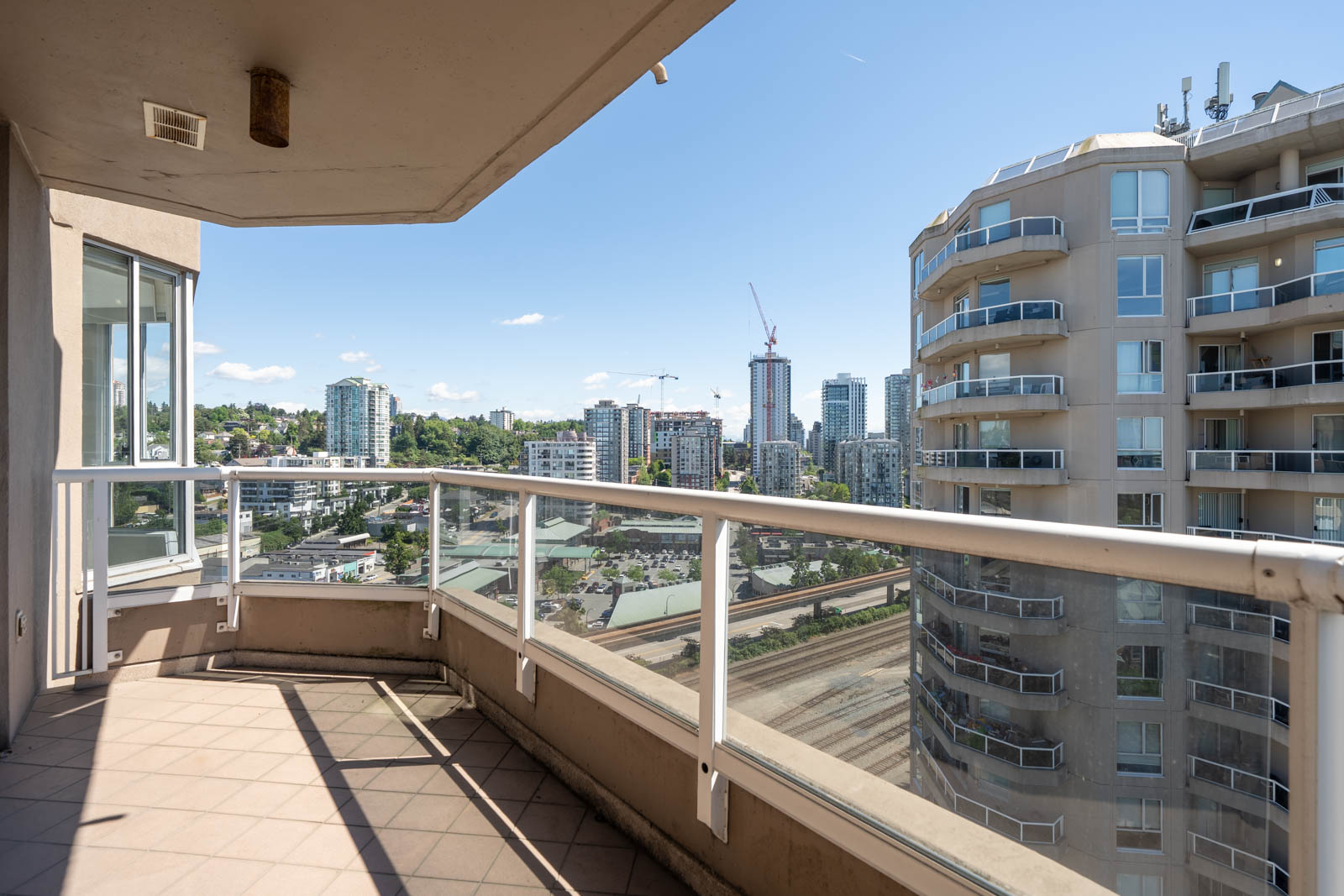View from a high-rise balcony overlooking nearby buildings, a construction crane, roads, and a clear sky in an urban setting.