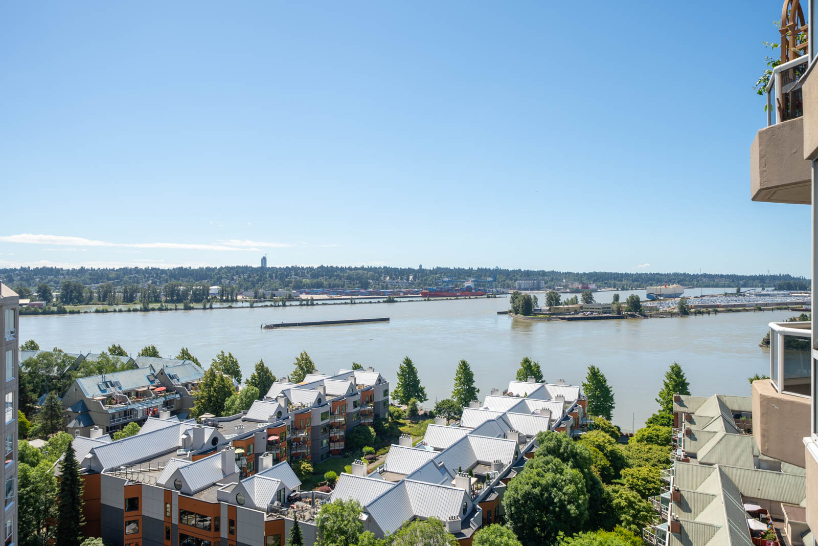 View from a high balcony overlooking residential buildings, trees, and a wide river with a barge; industrial buildings and a treeline are visible on the opposite bank under a clear sky.