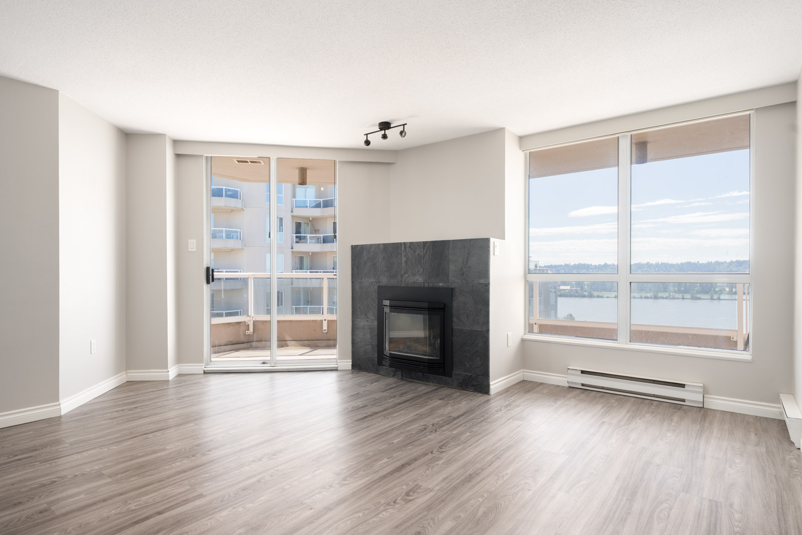 Unfurnished living room with large windows, a black fireplace, wood flooring, and a balcony door overlooking a river and nearby buildings.
