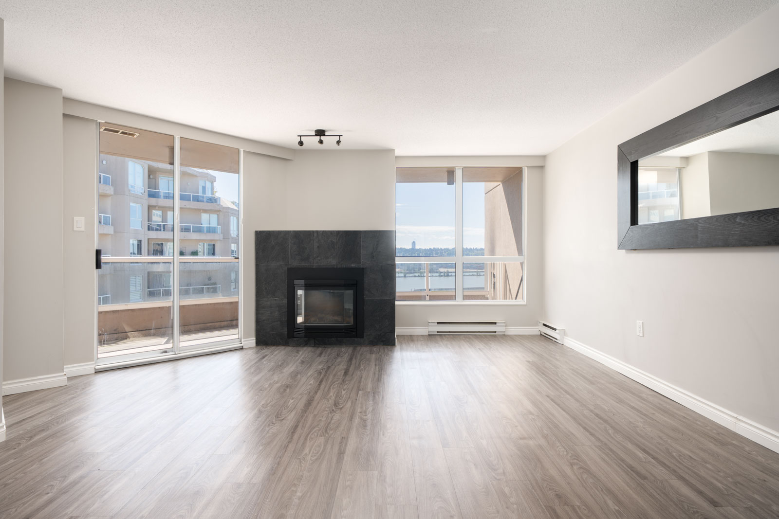 Unfurnished living room with wood flooring, a black fireplace, large windows, and a glass door opening to a balcony with a view of water and nearby buildings.