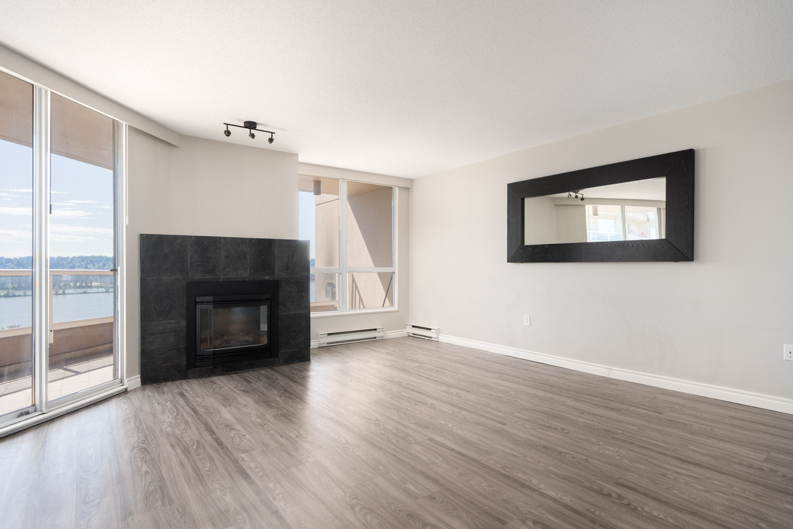 Empty living room with light gray walls, wood-look flooring, a black-framed fireplace, large windows, a wall mirror, and a balcony view of water and sky.