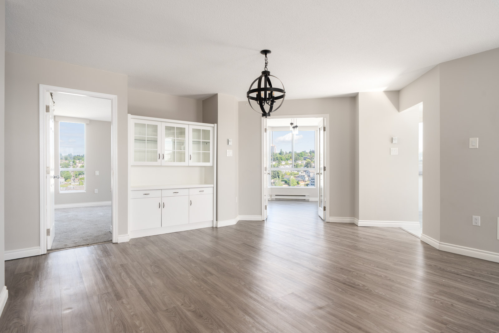 Bright, empty room with light wood flooring, built-in white cabinets, modern black chandelier, and large windows letting in natural light.