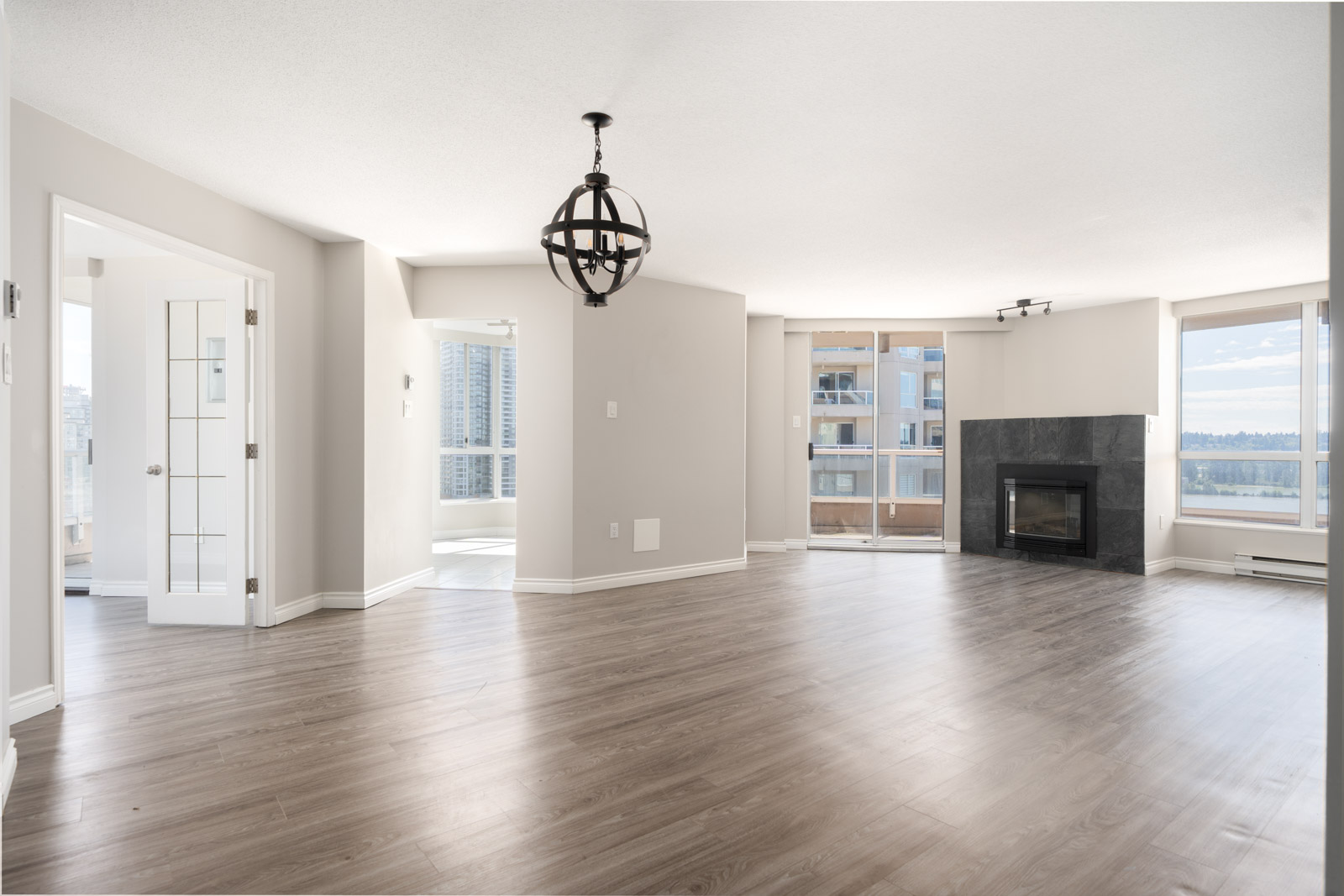 Spacious, empty living room with wood flooring, modern light fixture, large windows, glass doors to a balcony, and a fireplace. Natural light fills the space.