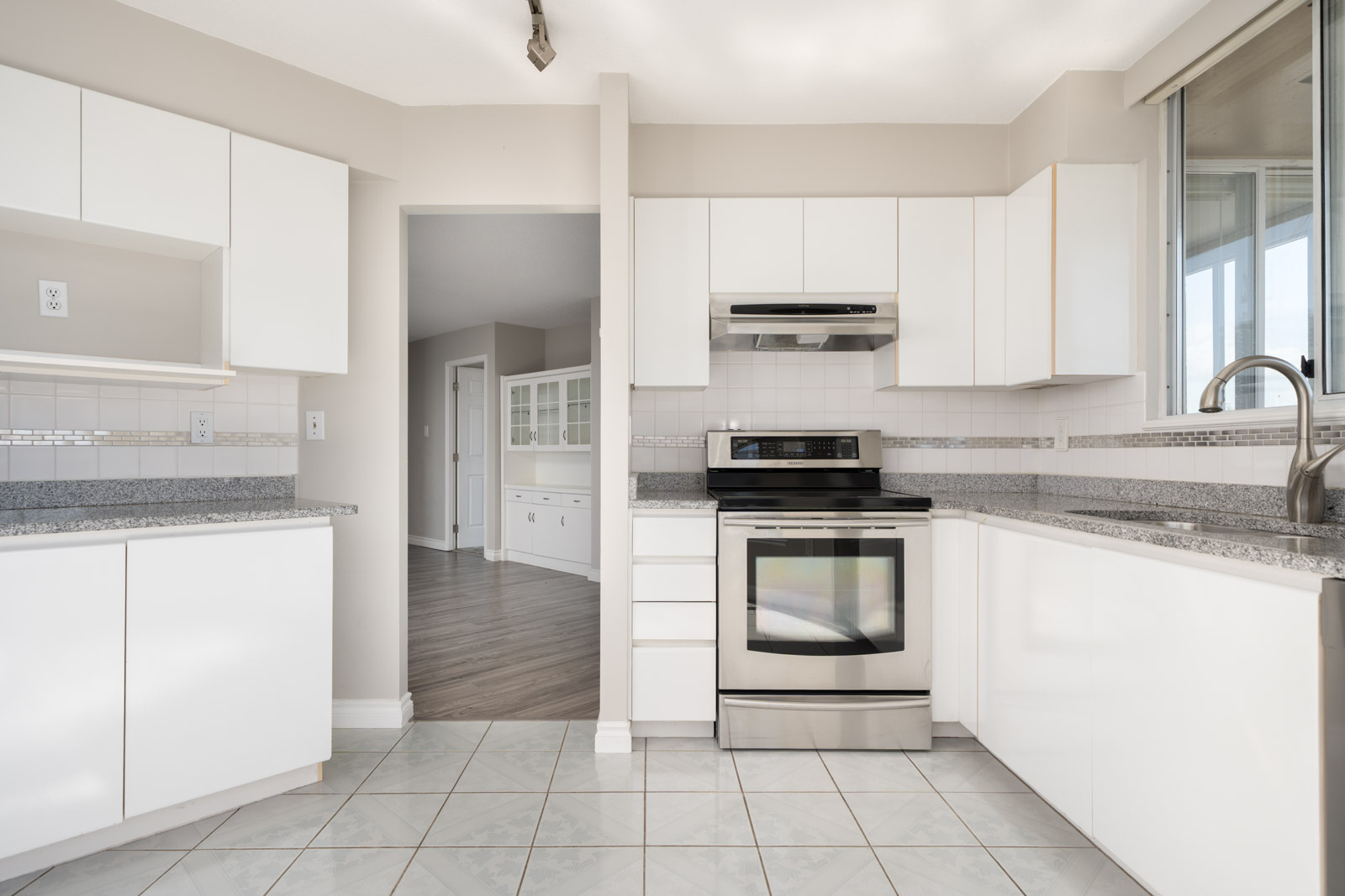 Modern kitchen with white cabinets, stainless steel stove and range hood, tile floor, granite countertops, and adjacent dining area visible through doorway.