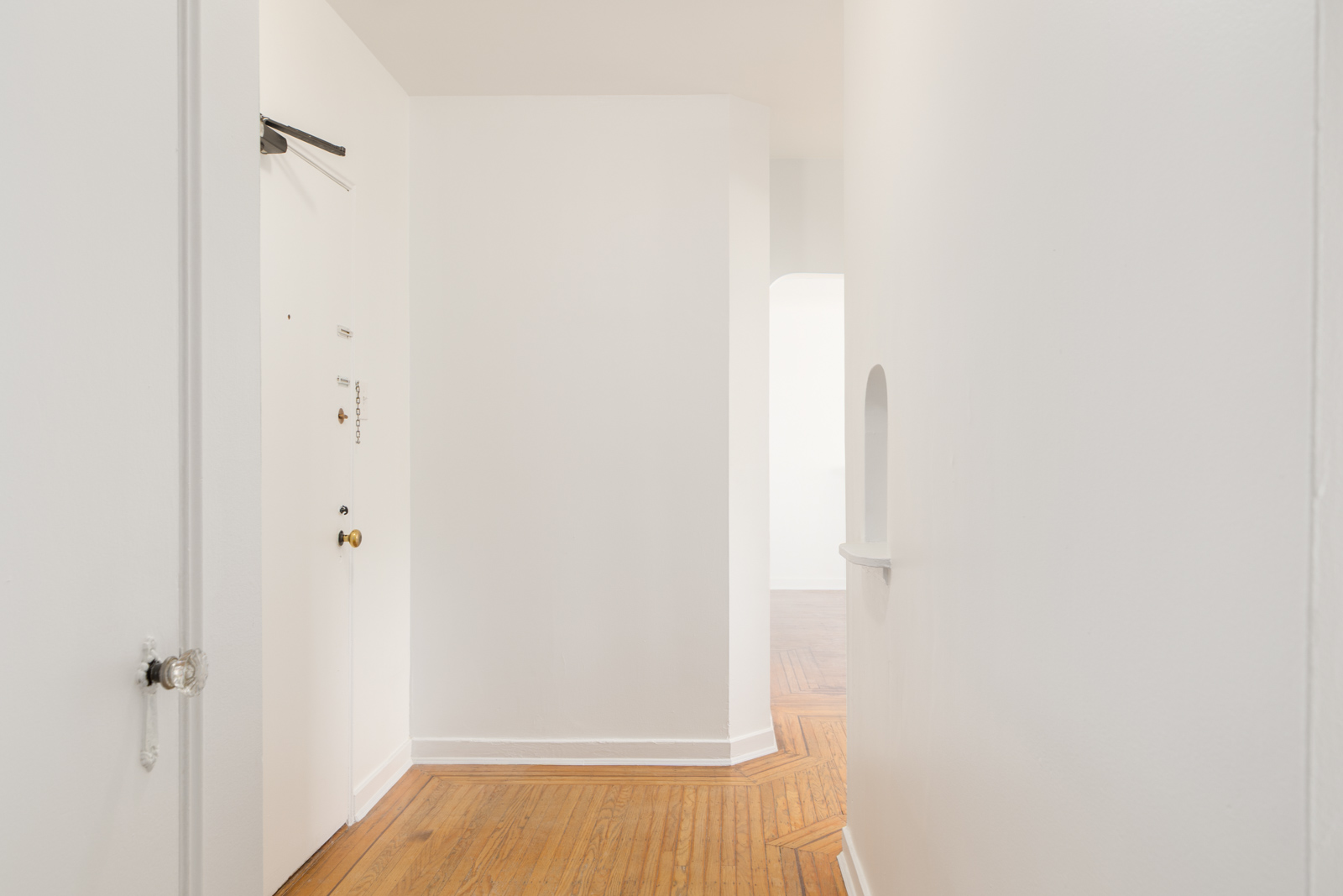 A white hallway with wooden floors, a closed white door on the left, and an arched wall opening leading to another room in the distance.
